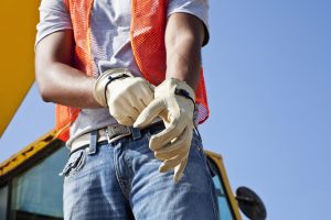 Low angle, cropped view of an African American young man putting on work gloves.  He is a construciton worker, wearing an orange safety vest and jeans.  Part of the crane is visible behind him, against the clear, blue sky.